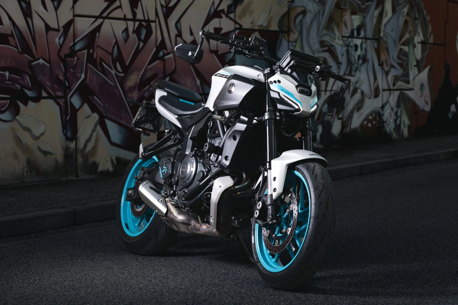 Yamaha MT-07 bianca con cerchi turchesi davanti a un muro di graffiti, ripresa a tre quarti